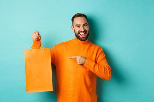 Satisfied male customer pointing at orange shopping bag, recommending store, smiling pleased, standing over turquoise background.