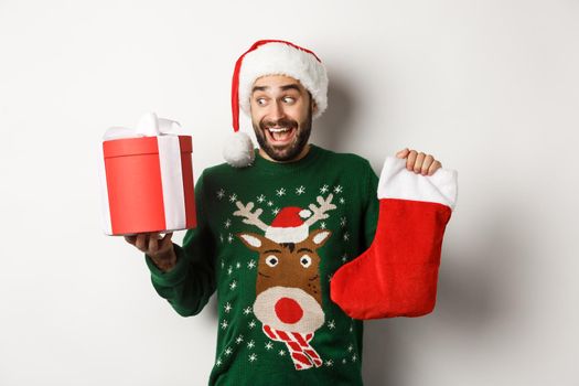 Xmas and winter holidays concept. Happy man got gifts on New Year eve, holding Christmas sock and present inside a box, standing over white background.