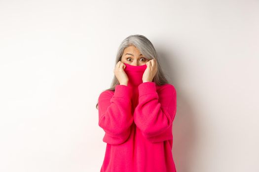 Portrait of funny asian grandmother hiding face in sweater collar, peeking at camera silly, standing over white background.