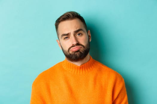 Close-up of upset young man sulking, looking disappointed and distressed, standing in orange sweater against turquoise background.