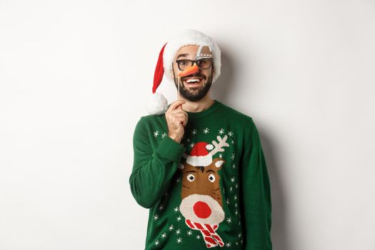 Christmas and holidays concept. Funny young man holding party mask and wearing xmas Santa hat, smiling happy, celebrating new year, white background.