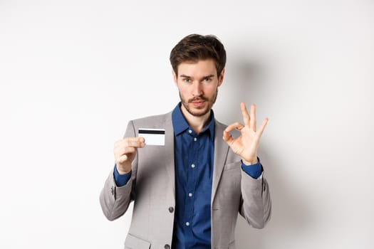 Happy male bank client in business suit showing plastic credit card and okay sign, smiling satisfied, standing on white background.