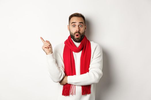 Christmas shopping and winter holidays concept. Surprised bearded guy checking out new year discounts, pointing finger left at empty space, standing over white background.