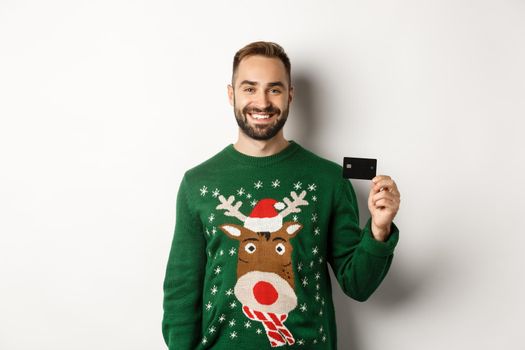 Christmas holidays and shopping concept. Young man in winter sweater showing credit card, standing over white background.
