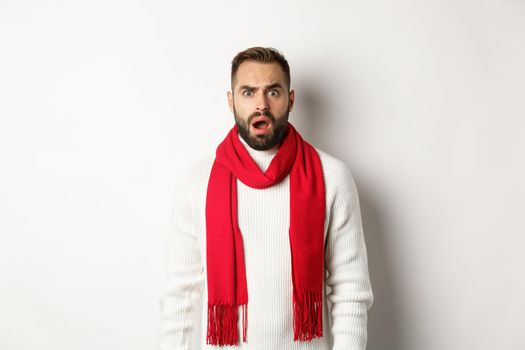 Christmas holidays and celebration concept. Confused bearded guy staring at something strange, standing in red scarf and sweater, white background.
