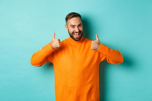 Handsome man expressing support, showing thumb up, encourage you, praise excellent work, approve and agree, standing over light blue background.