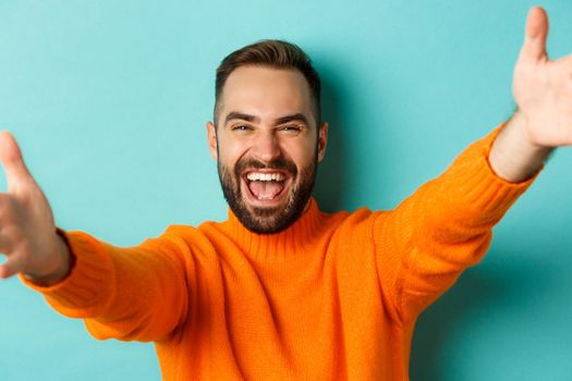 Close-up of handsome happy man reaching hands forward, stretching arms for hug, standing against turquoise background.