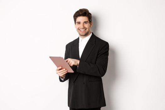 Image of handsome businessman in black suit, using digital tablet and smiling, standing against white background.