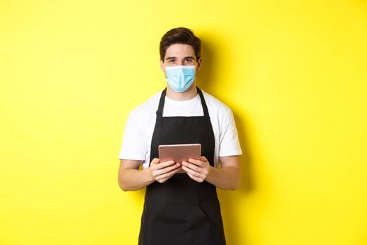 Concept of covid-19, small business and pandemic. Waiter in black apron and medical mask taking order, holding digital tablet, standing over yellow background.