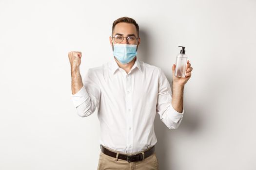 Covid-19, social distancing and quarantine concept. Cheerful manager in medical mask showing hand sanitizer, standing over white background.