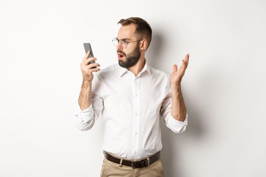 Man looking confused at mobile phone after conversation, standing puzzled over white background. Copy space