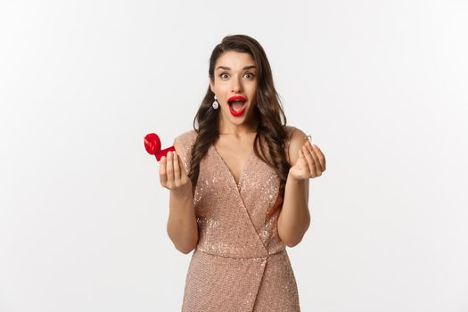 Excited woman holding engagement ring and red box, looking surprised and amazed, receive marriage proposal on date, wearing elegant dress, standing over white background.