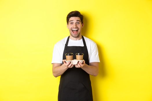 Excited male barista holding two cups of takeaway coffee, working in cafe, standing over yellow background.