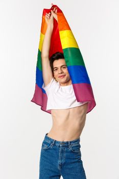 Vertical view of beautiful androgynous gay man raising rainbow flag and smiling happy, standing in crop top and jeans against white background.
