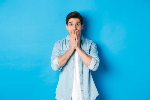 Portrait of shocked adult man looking at camera in awe, gasping and covering mouth with hands, standing over blue background.