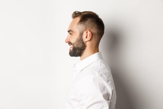 Close-up profile shot of handsome bearded man looking left and smiling, standing against white background.