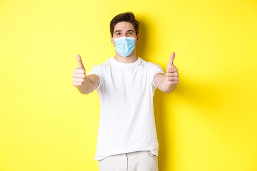 Concept of coronavirus, pandemic and social distancing. Confident man protecting himself from covid-19 with medical mask, showing thumbs up in approval, yellow background.
