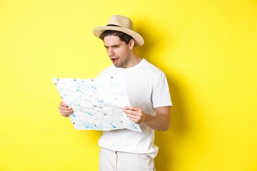 Travelling, vacation and tourism concept. Tourist looking displeased and shocked at road map, standing against yellow background.