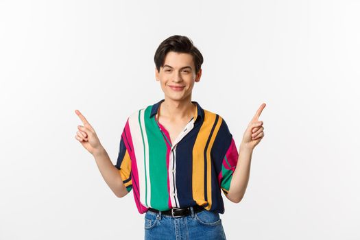 Handsome queer man pointing fingers sideways, smiling pleased and showing two choices, standing over white background.