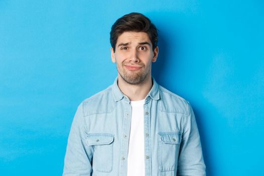 Close-up of skeptical and awkward guy smirking, feeling uncomfortable, standing over blue background.