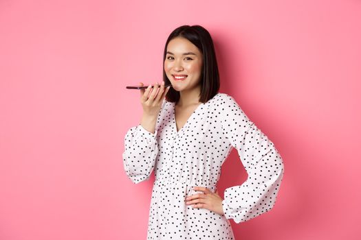 Beautiful korean woman talking on speakerphone, recording voice message and smiling happy, standing over pink background.