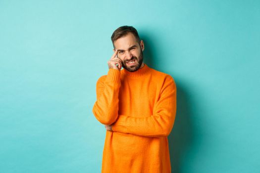 Man with beard, grimacing and cringe from something embarrassing, looking with awkward face, standing over light blue background.