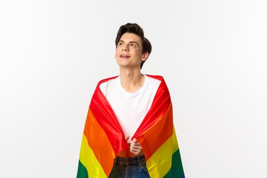 Human rights and lgbtq community concept. Beautiful young androgynous man with glitter on face, wearing rainbow pride flag and looking at upper left corner, white background.