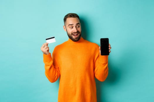 Online shopping. Amazed guy using credit card and showing mobile screen, looking impressed, standing against light blue background.