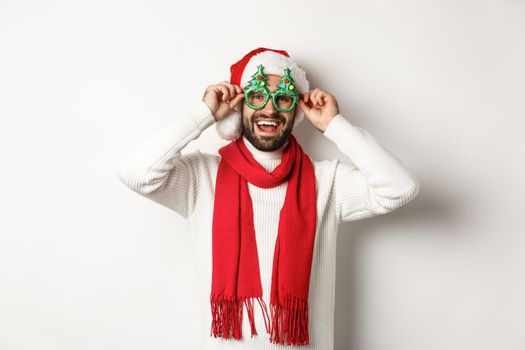 Christmas, New Year and celebration concept. Happy man laughing, wearing Santa hat and party glasses, standing over white background.