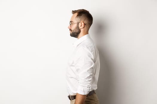 Profile of young businessman in white collar shirt and beige pants, looking left, standing against studio background.