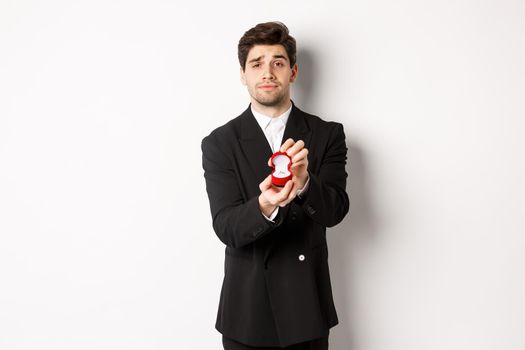 Portrait of handsome man making a proposal, asking to marry him, showing ring, standing in black suit against white background.