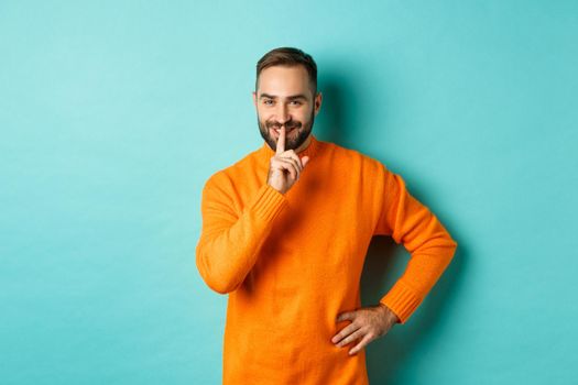 Happy man asking keep voice down, shushing and smiling, prepare surprise, hushing at camera, standing over turquoise background.