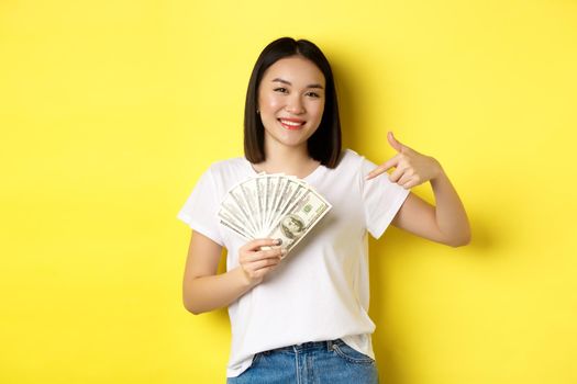 Young asian woman smiling, showing prize money, pointing finger at dollars, standing over yellow background.