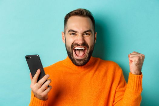Image of man winning on mobile phone, making fist pump and rejoicing of victory, holding smartphone with satisfied face, turquoise background.