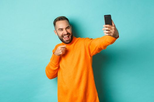 Handsome man video calling on mobile phone, taking selfie and pointing at smartphone camera, standing over turquoise background.