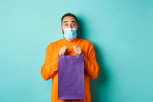 Covid-19, pandemic and lifestyle concept. Surprised man open shopping bag and looking amazed, receiving gift on holiday, standing over turquoise background.