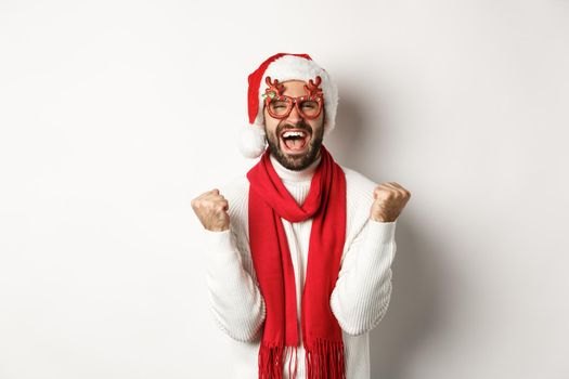 Christmas, New Year and celebration concept. Excited man in Santa hat and party glasses, winning and rejoicing, making fist pump, shouting yes, standing over white background.