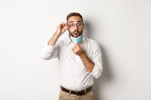 Covid-19, social distancing and quarantine concept. Impressed business man take off medical mask, looking surprised, standing over white background.