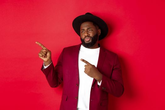 Winter holidays and shopping concept. Skeptical and displeased Black man looking with disdain and pointing finger left at copy space, standing against red background.