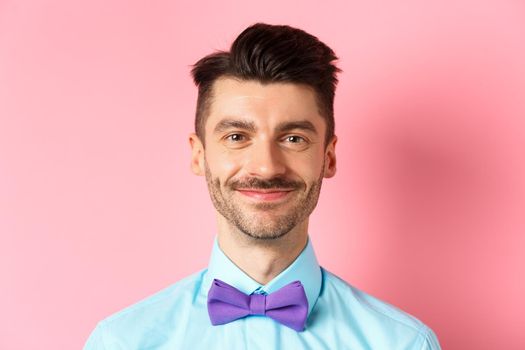 Close-up of smiling hansome man with moustache, wearing bow-tie and shirt, standing cheerful on pink background.