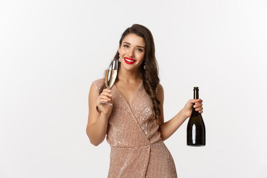 Winter holidays celebration concept. Image of happy young woman in luxury dress, drinking champagne from glass, holding bottle, New Year party, white background.