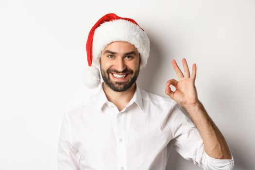 Party, winter holidays and celebration concept. Confident man in santa hat showing okay sign, approve and like, white background.