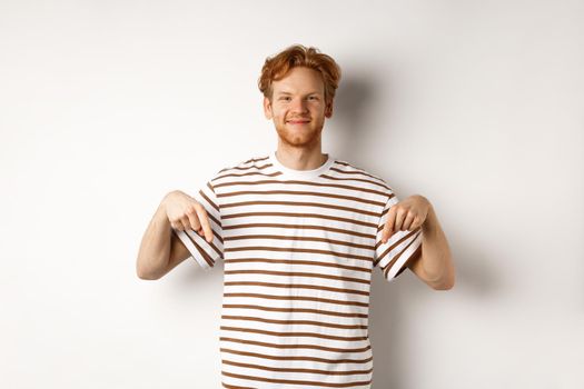 Handsome male model with ginger hair and bristle, pointing fingers down and smiling, showing advertisement, white background.