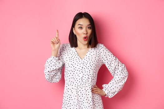 Excited asian girl having an idea, saying suggestion, raising finger and talking, standing over pink background.