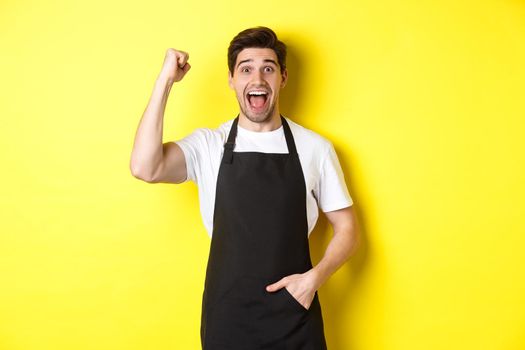 Cheerful seller making fist pump, rejoicing and triumphing, standing in black apron against yellow background.
