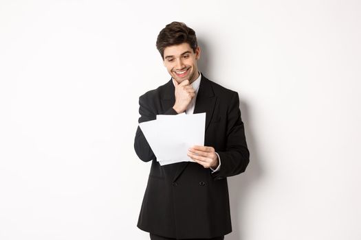 Image of handsome businessman in black suit, looking pleased at documents, reading report and smiling, standing against white background.