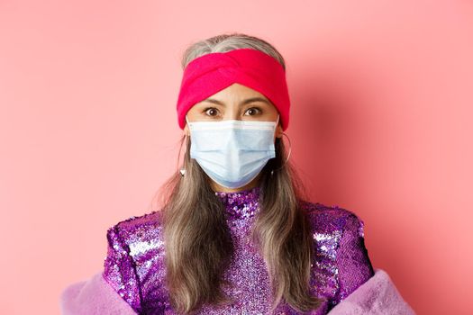 Covid-19, virus and social distancing concept. Close-up of stylish asian senior woman in medical mask and shiny dress looking at camera, standing over pink background.