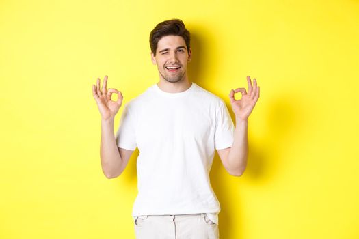 Confident handsome man winking, showing okay signs in approval, like something good, standing over yellow background.