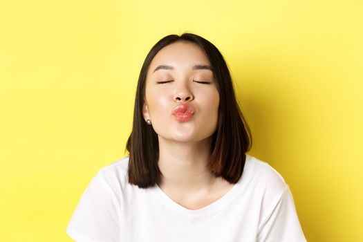 Valentines day concept. Close up of cute asian girl pucker lips and close eyes for kiss, standing romantic against yellow background.