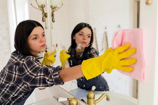 A young girl is cleaning the bathroom, applying detergent with a spray and washing the mirror with a sponge in yellow gloves on her hands. Smiling woman taking care of the cleanliness of her home.